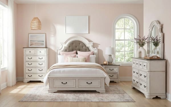 Bedroom setup with white wooden furniture including a tufted headboard bed, nightstand, tall dresser, and wide dresser with mirror, soft pink walls, and natural light.